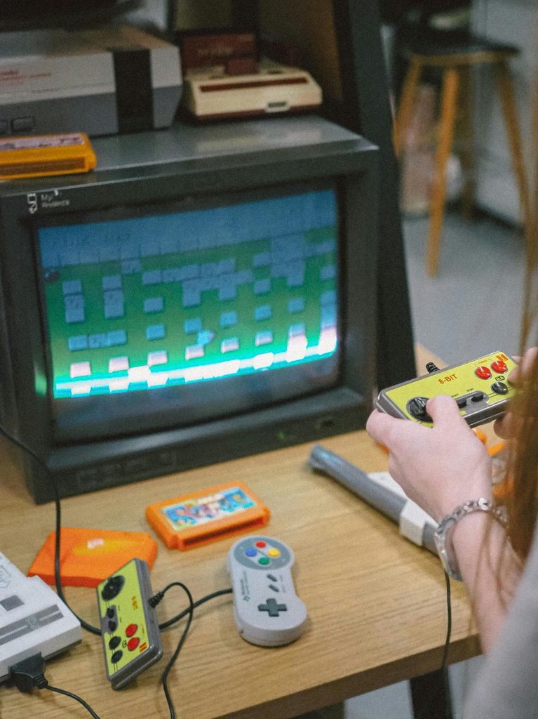 Person hält einen gelben 8-Bit-Controller, auf dem Tisch liegen weitere Retro-Controller und Spielmodule vor einem CRT-Bildschirm